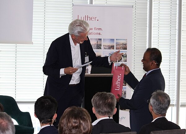A man in a suit hands a gift bag labeled "Luther" to another man, who is also in formal attire. The setting appears to be a professional event, with seated audience members observing the exchange.