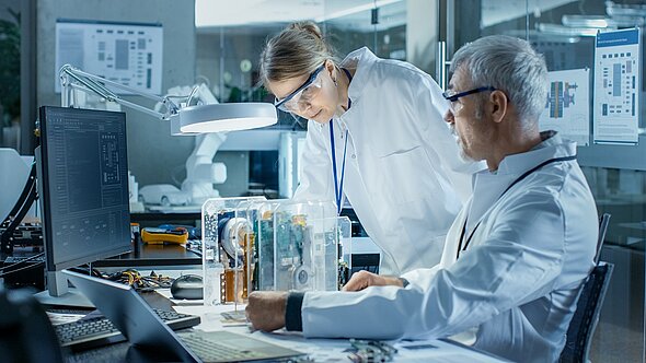 Two scientists in lab coats examine a transparent device on a table in a modern laboratory. One is leaning over the device, while the other is focused on a computer screen. The workspace is well-lit, with tools and equipment visible.