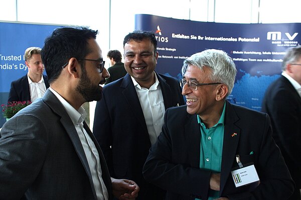 A group of three men engage in conversation, smiling and looking at each other. One man is wearing glasses and a suit, while another has light hair and a casual shirt. The background features a large banner with text, suggesting a professional or networking event.