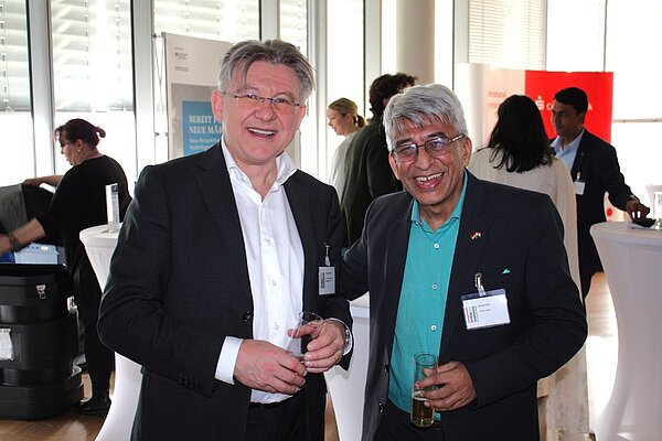 Two smiling men stand together at an event, holding drinks. One man has gray hair and glasses, wearing a dark suit over a light shirt. The other has silver hair and is dressed in a black jacket over a turquoise shirt. Background figures are engaged in conversations.