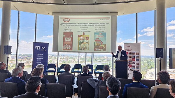 A speaker presents during a conference, discussing consumer-centric strategies. A projection behind him displays product images, including milk, mayonnaise, and peanut butter, highlighting their benefits. The audience, visible in the foreground, listens attentively, with a cityscape visible through large windows in the background.