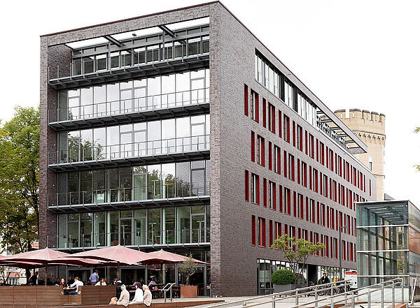 A modern multi-story building features large glass windows and red accents. In the foreground, a sloped accessible ramp leads to the entrance. Several people sit at outdoor tables under umbrellas, creating a lively atmosphere. A bicycle is parked nearby.