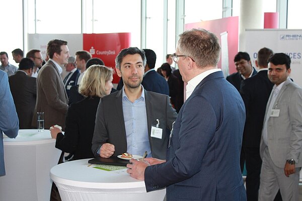 A group of professionals engages in conversation at a networking event. Two men in business attire stand at the forefront, one holding a small plate and the other with a notepad. The atmosphere is lively, with people talking in the background.