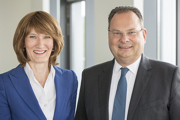 A woman with shoulder-length brown hair, wearing a blue blazer, smiles alongside a man with short hair, wearing glasses and a dark suit. They are positioned in a bright, modern indoor setting, conveying a professional and friendly atmosphere.