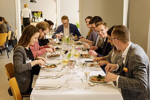 Eine Gruppe von elf Personen sitzt an einem elegant gedeckten Tisch und genießt ein gemeinsames Abendessen. Es gibt verschiedene Gerichte auf den Tellern, und die Gäste unterhalten sich fröhlich, während sie essen und trinken. Der Raum hat eine warme, einladende Atmosphäre.