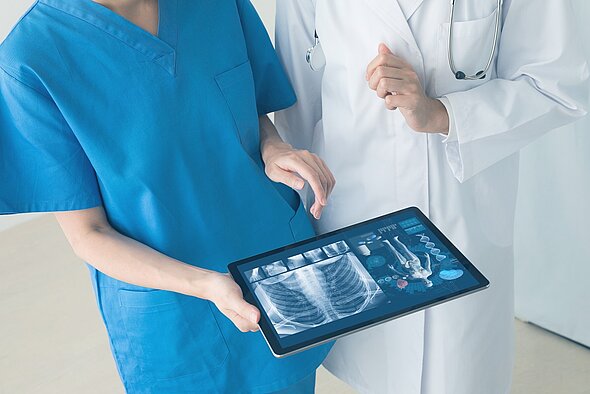 Two medical professionals are discussing a digital tablet displaying X-ray images and health data. One is wearing blue scrubs, while the other is in a white coat. They are closely examining the information on the tablet, indicating a collaborative diagnostic process.