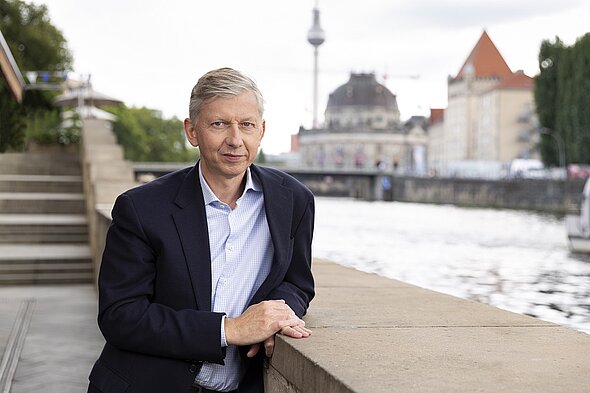 Ein Mann in einem dunklen Blazer steht an einem Steinpfeiler am Flussufer. Er lächelt und schaut in die Kamera. Im Hintergrund sind Gebäude und ein Turm sichtbar. Es ist ein klarer Tag mit teilweise bewölktem Himmel.