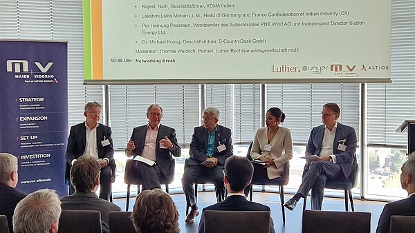 A panel discussion featuring five speakers seated and engaging with the audience. They represent various organizations and industries, discussing topics related to business and international cooperation. The setting includes large windows, modern furnishings, and branding elements in the background.