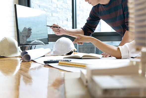 Two individuals collaborate at a table covered with blueprints, an open notebook, and a calculator. One person points to a computer screen displaying a project plan, while the other gestures towards the screen, engaged in discussion. Hard hats are visible nearby.