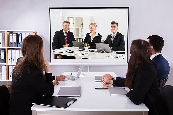 In einem modernen Konferenzraum sitzen fünf Personen. Vier schauen auf einen großen Bildschirm, auf dem drei Personen in Anzügen an einem Tisch stehen. Es gibt Laptops und Unterlagen auf dem Tisch, sowie Stifte und einen Blumentopf im Hintergrund.