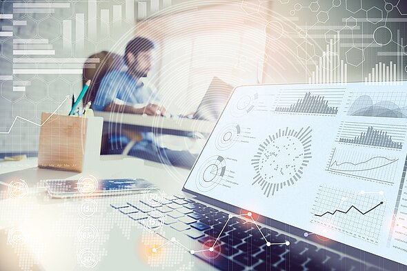 Ein Laptop mit Diagrammen und Grafiken steht im Vordergrund. Die Grafiken zeigen verschiedene Datenanalysen und Statistiken. Im Hintergrund ist eine Person zu sehen, die an einem Tisch arbeitet. Die Atmosphäre ist modern und technologisch.