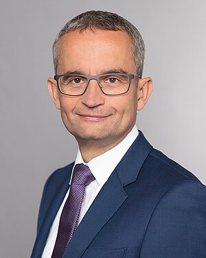 Das Bild zeigt einen Mann mit kurzem, grauem Haar und Brille, der in einem blauen Anzug mit weißem Hemd und lila Krawatte gekleidet ist. Er lächelt leicht und blickt direkt in die Kamera, vor einem grauen Hintergrund.