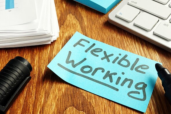 Ein blauer Haftnotizzettel liegt auf einem Holzschreibtisch und trägt die Aufschrift „Flexible Working“. Daneben befinden sich ein Stapel Papier und eine Tastatur. Ein schwarzer Stift ist ebenfalls sichtbar.
