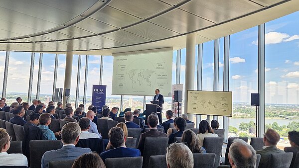 A speaker presents to an audience in a modern conference room with large windows. Behind the speaker is a screen displaying data titled "MR at a glance," showing statistics and a world map. The audience, dressed in business attire, is engaged with the presentation.