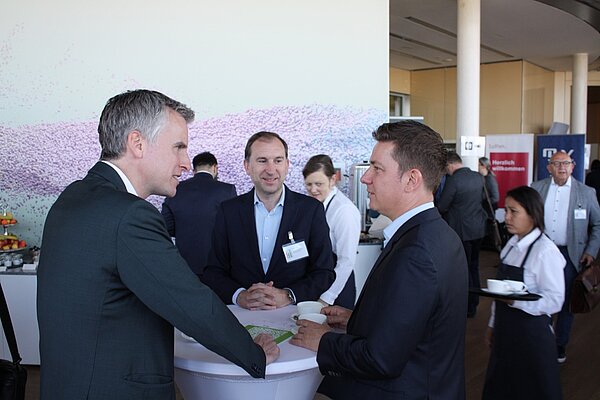 A group of four people engaged in conversation at a networking event. Two men are interacting at a table, while a third man listens. In the background, a woman in a uniform carries a tray, and other attendees are mingling. The atmosphere is professional and social.