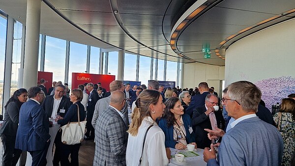 A large group of people are engaged in conversation at a networking event held in a modern, glass-walled venue. Some attendees are holding drinks, while others are standing at tables. The atmosphere appears professional and lively, with various banners visible in the background.