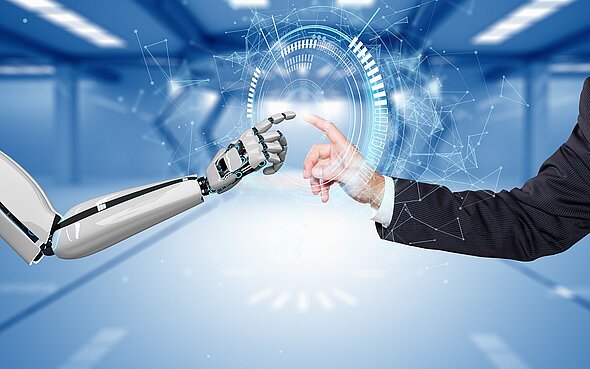 A robotic arm and a human hand are reaching towards each other, almost touching. Digital elements and glowing circular graphic overlays are surrounding the connection point, symbolizing interaction between technology and humanity. The background features a blurred, futuristic setting.