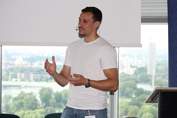 A man wearing a white t-shirt gestures while speaking at an event. He stands in front of a large window, displaying a view of green trees and buildings in the distance. The image conveys an engaging presentation or discussion taking place.