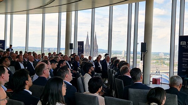 A large audience of professionals sits in a modern conference room with floor-to-ceiling windows, overlooking a cityscape. The background features a historic cathedral with distinctive spires. Attendees listen attentively to a speaker, engaged in a business presentation.