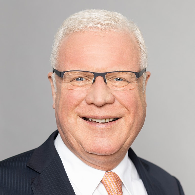 Ein älterer Mann mit grauen Haaren trägt eine Brille und einen dunklen Anzug mit orangefarbener Krawatte. Er lächelt freundlich in die Kamera. Der Hintergrund ist grau.