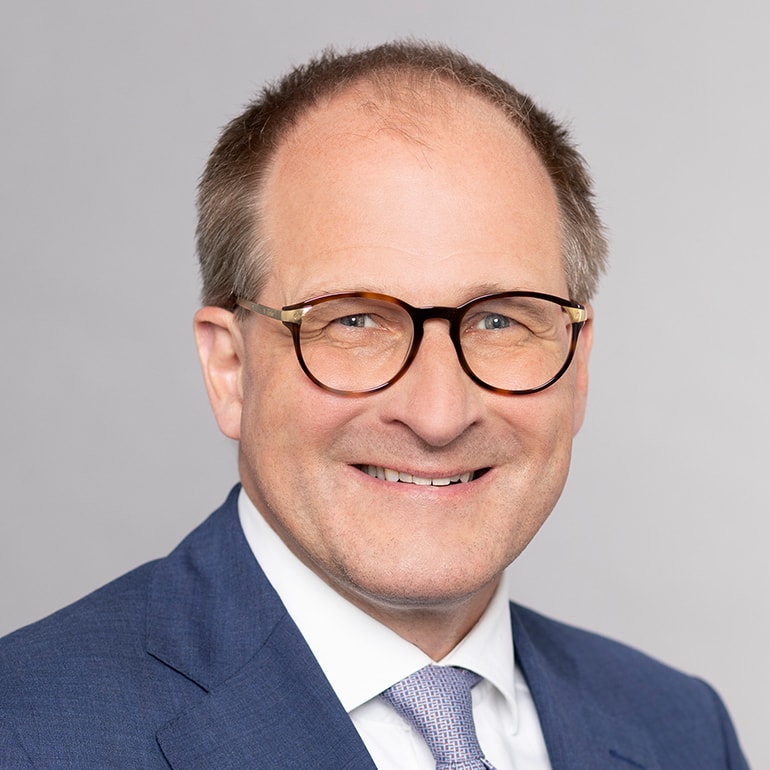 A smiling man with short, light hair is wearing round glasses and a blue suit with a white shirt and patterned tie. The background is light gray, emphasizing his professional appearance.