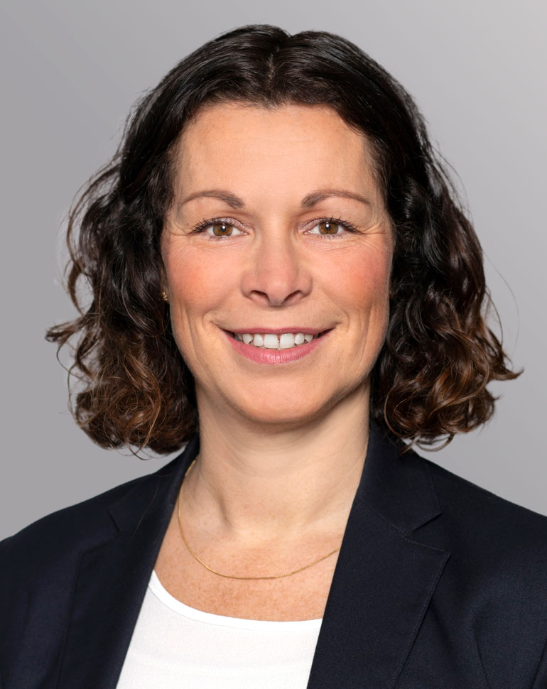 Porträt einer lächelnden Frau mit lockigem, braunem Haar, die einen dunklen Blazer über einem weißen Oberteil trägt. Der Hintergrund ist grau. Die Frau wirkt freundlich und professionell.