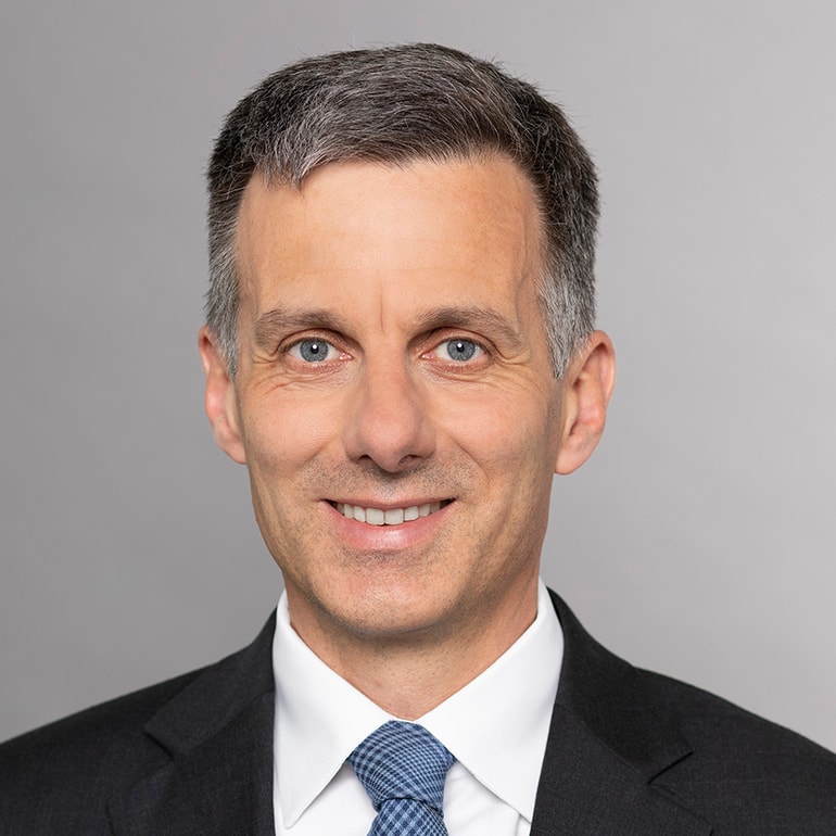 A smiling man with short, light gray hair and blue eyes is wearing a dark suit, white shirt, and a blue tie. The background is a neutral gray color.