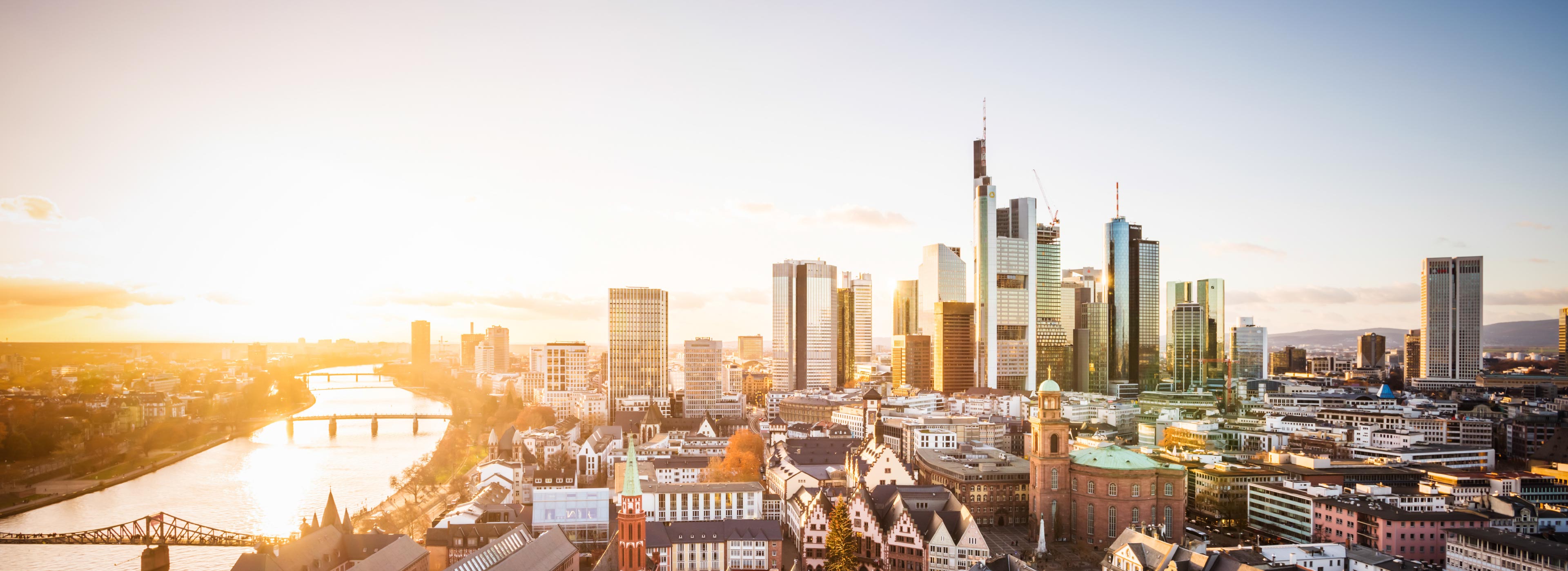 Stadtansicht von Frankfurt am Main bei Sonnenuntergang. Moderne Hochhäuser dominieren die Skyline, während der Mainfluss im Vordergrund verläuft. Die Architektur vereint historische und zeitgenössische Gebäude, die eine lebendige städtische Atmosphäre schaffen.
