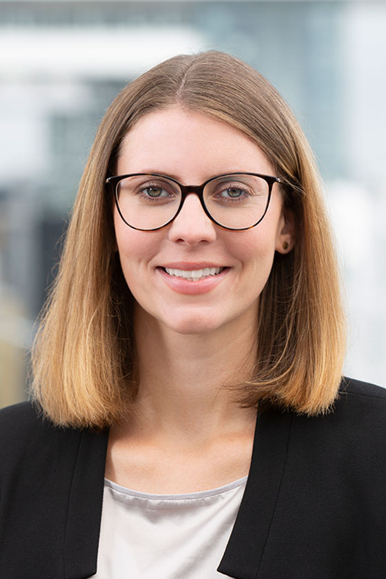 Porträt einer Frau mit langen, hellbraunen Haaren und einer Brille. Sie trägt einen schwarzen Blazer und lächelt freundlich. Der Hintergrund ist unscharf und zeigt eine moderne Gebäudeansicht.