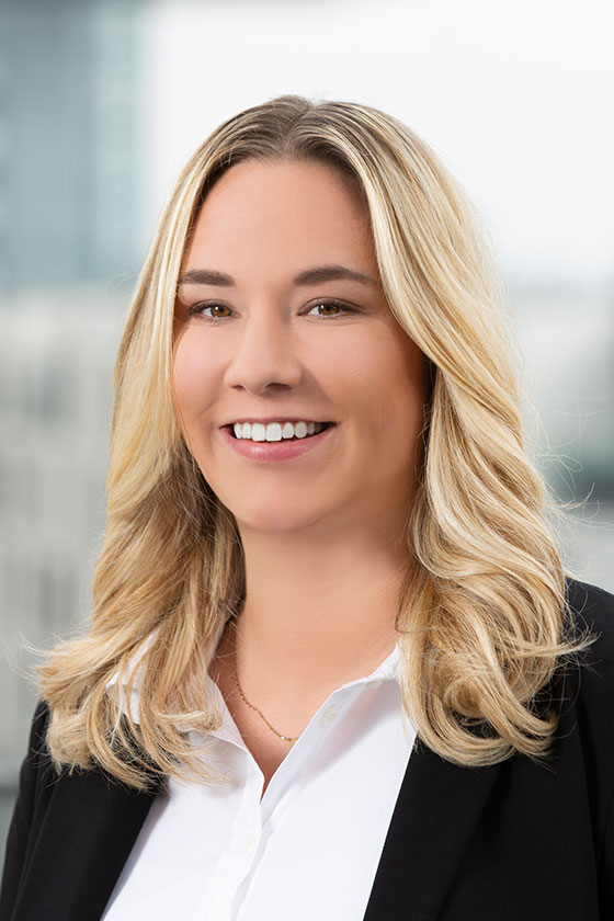 Eine lächelnde Person mit langen, blonden Haaren trägt ein schwarzes Blazerjackett und ein weißes Hemd. Der Hintergrund ist unscharf und zeigt eine moderne Büroumgebung.