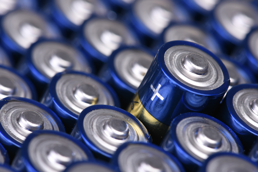 Eine Ansammlung von blauen Batterien, die dicht nebeneinander liegen. Eine Batterie hebt sich hervor, mit einem Plus-Zeichen auf der oberen Kappe. Die Oberfläche der Batterien ist glänzend und reflektiert Licht.