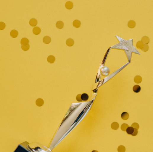 Ein silberner Pokal mit einer stilisierten Figur, die einen Stern hält, steht auf einem schwarzen Sockel. Der Hintergrund ist gelb mit goldenen Konfetti-Teilchen, die eine festliche Atmosphäre schaffen.