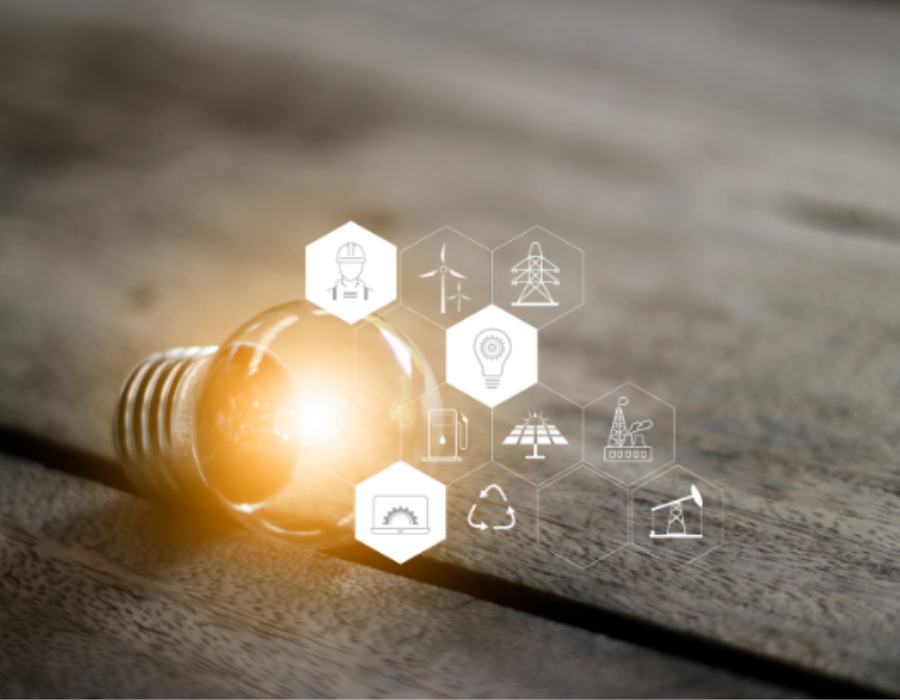 Eine Glühbirne auf einem Holzuntergrund, umgeben von Hexagon-Symbolen, die verschiedene Energiequellen und Nachhaltigkeit darstellen, wie Windkraft, Solarenergie und Recycling. Die Glühbirne leuchtet, was den Fokus auf Energieeffizienz und innovative Technologien legt.