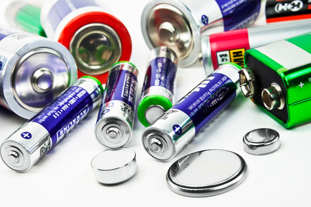 Eine Sammlung von Batterien unterschiedlicher Größe und Typen, einschließlich AA-Batterien, Knopfzellen und einer 9-Volt-Batterie. Die Batterien liegen auf einem weißen Hintergrund und sind in verschiedenen Farben, hauptsächlich Silber und Schwarz, mit klaren Plus- und Minuszeichen.