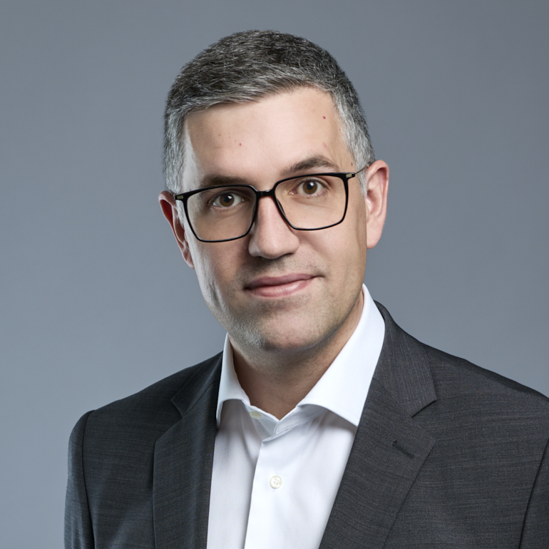 Porträt eines Mannes mit kurzen grauen Haaren, der eine Brille trägt. Er trägt ein dunkles Sakko und ein weißes Hemd. Der Hintergrund ist hellgrau. Der Mann schaut direkt in die Kamera und hat einen neutralen Gesichtsausdruck.