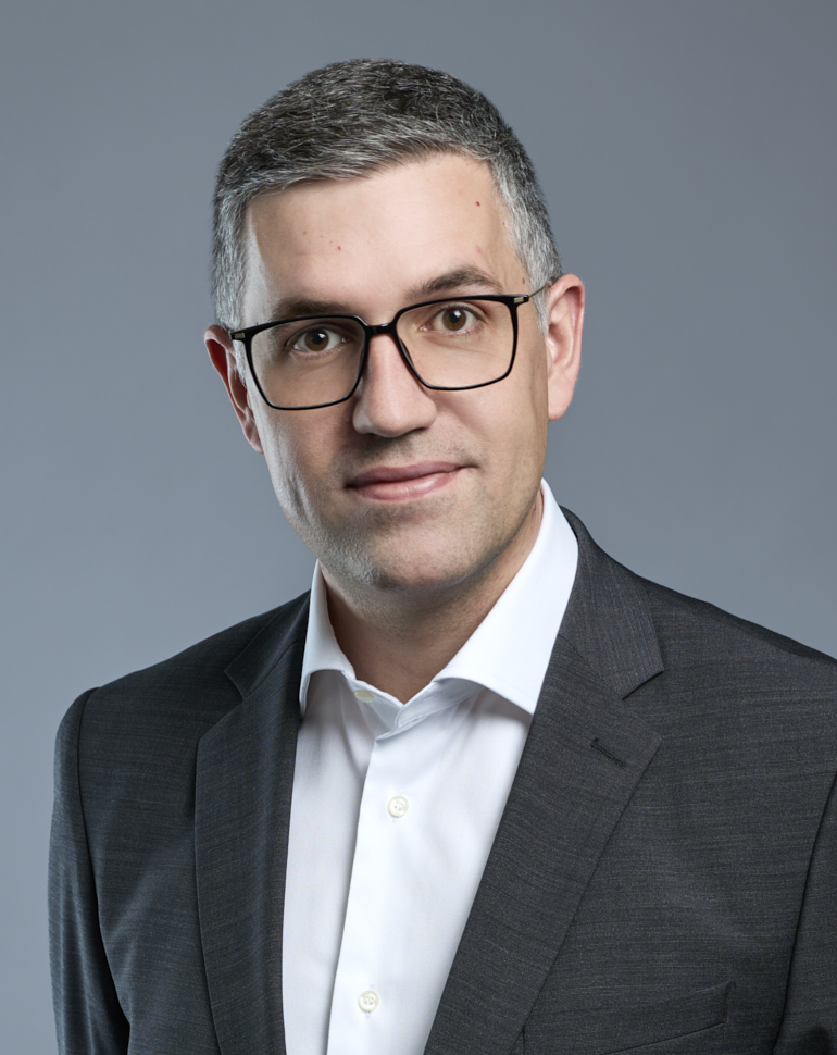 Porträt eines Mannes mit kurzen, grauen Haaren, der eine Brille trägt. Er ist in einem dunklen Anzug und einem weißen Hemd gekleidet. Der Hintergrund ist einfarbig grau. Der Mann schaut direkt in die Kamera und wirkt freundlich.