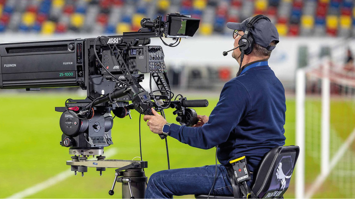 Ein Kameramann sitzt auf einem Stuhl und bedient eine große Fernsehkamera, die auf ein Spielfeld ausgerichtet ist. Er trägt Kopfhörer und spricht in ein Mikrofon, während im Hintergrund unklare farbige Sitzplätze zu sehen sind.