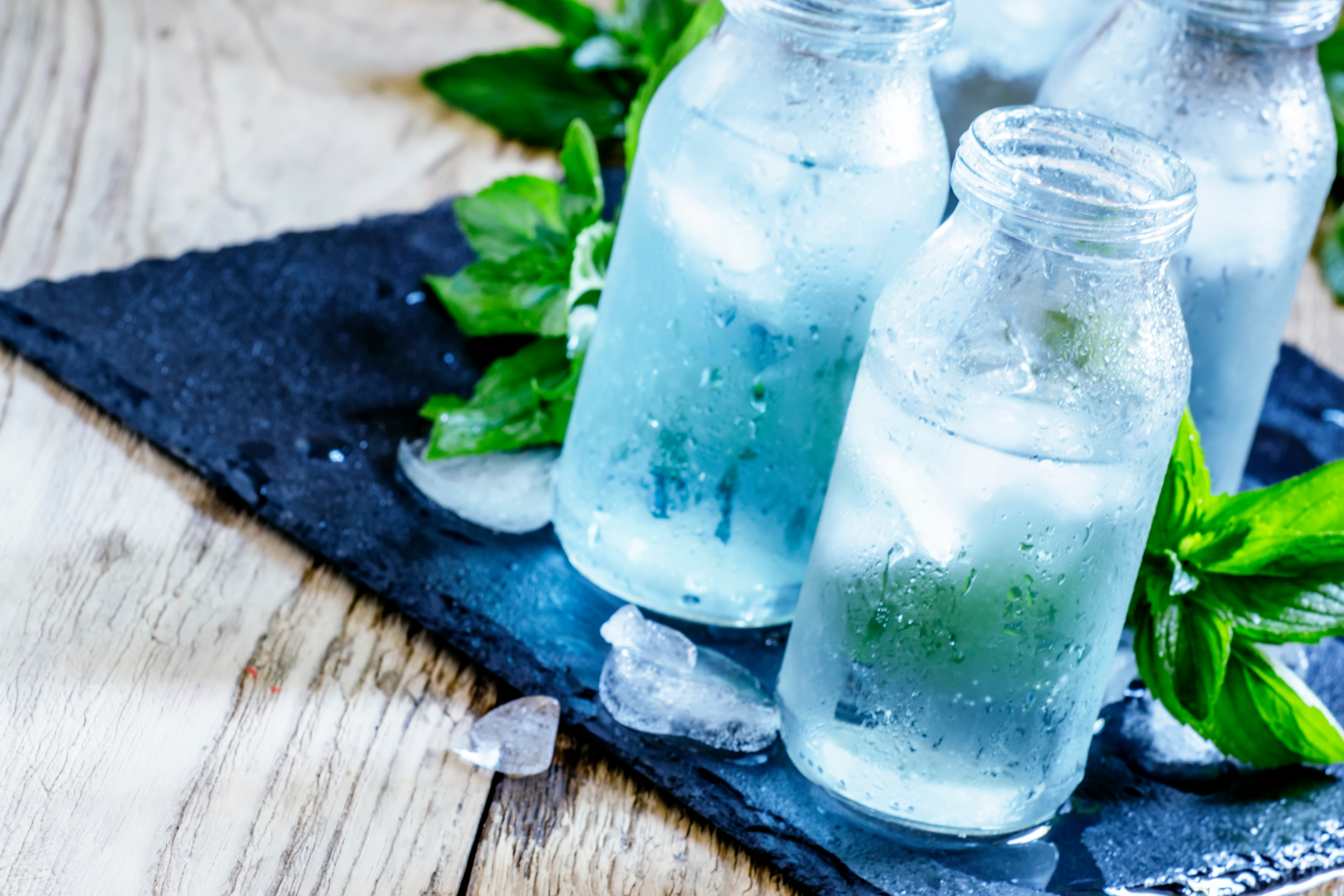 Four glass bottles filled with cold, clear water are placed on a slate board. Ice cubes and fresh mint leaves surround the bottles, emphasizing a refreshing beverage aesthetic. The condensation on the glass suggests the coolness of the drinks.