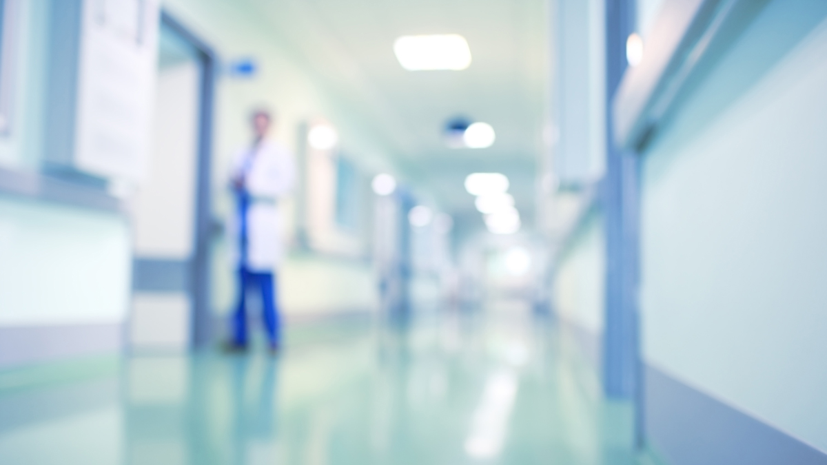 A blurred hospital corridor is depicted, featuring a healthcare professional wearing a white coat and blue pants, standing in the distance. The environment suggests a clinical setting with soft lighting and greenish floor tiles, indicating the atmosphere of a medical facility.