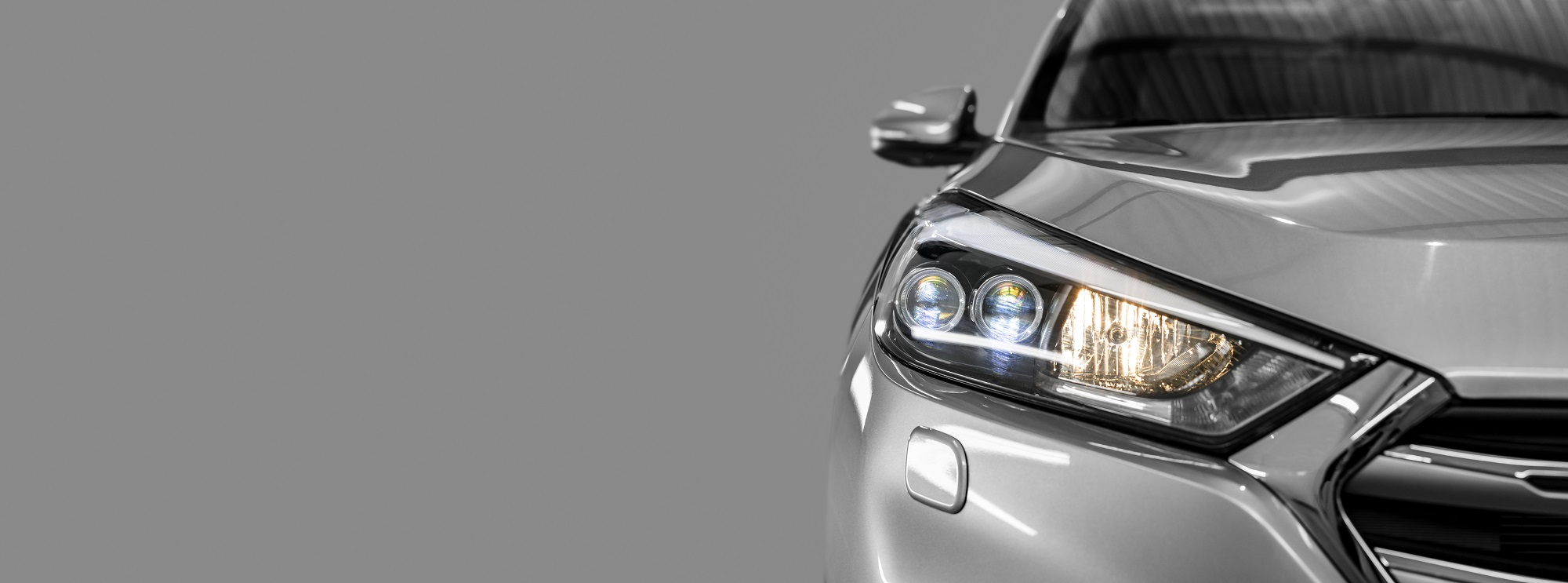 Close-up of a sleek silver car's front end, focusing on the headlights and grille. The left headlight features bright LED lights, enhancing the car's modern design. The background is a neutral gray, emphasizing the car's polished finish.