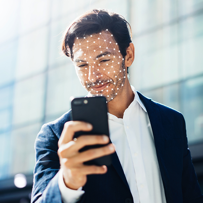 Ein Mann in einem Anzug hält ein Smartphone und schaut lächelnd auf den Bildschirm. Auf seinem Gesicht sind geometrische Linien und Punkte dargestellt, die eine digitale Analyse seines Gesichts symbolisieren, möglicherweise für Gesichtserkennungstechnologie. Im Hintergrund ist eine unscharfe moderne Gebäudefassade sichtbar.