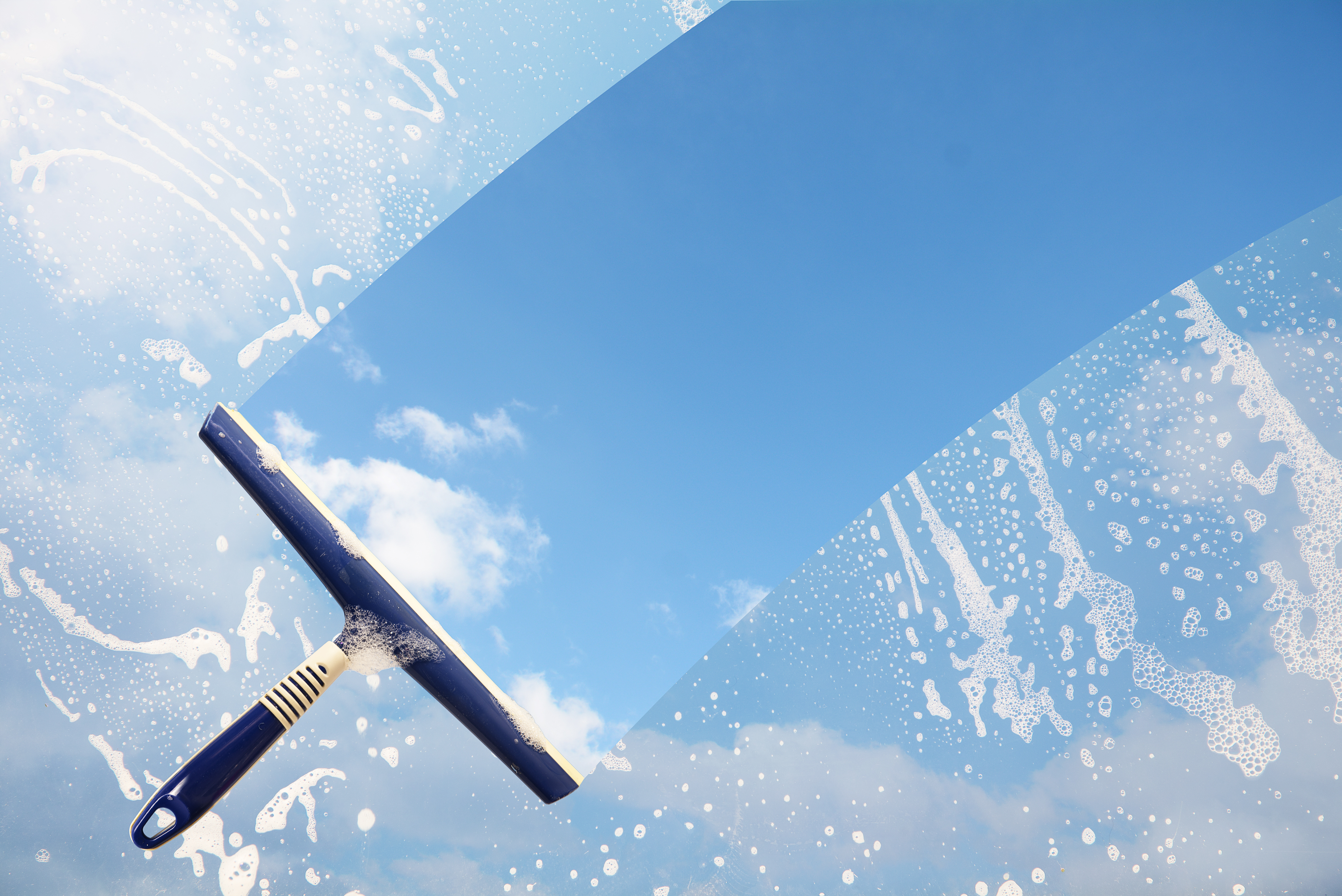 A squeegee is positioned against a bright blue sky. The area around it shows soap bubbles and streaks, indicating a cleaning process. The contrast illustrates a clear section amidst the soapy surface, symbolizing clarity and freshness.