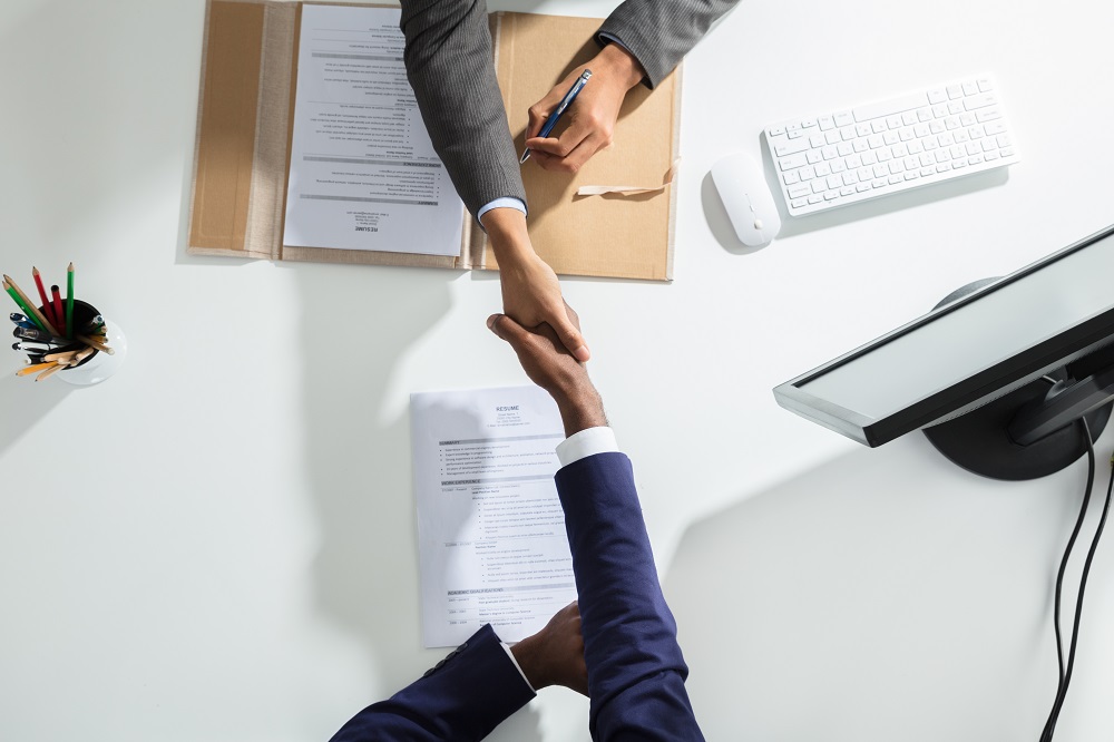 Two people are shaking hands across a table, suggesting a successful agreement or partnership. One individual is holding a pen, and there are documents, including a resume, visible on the table. A keyboard and a monitor are also present in the background.