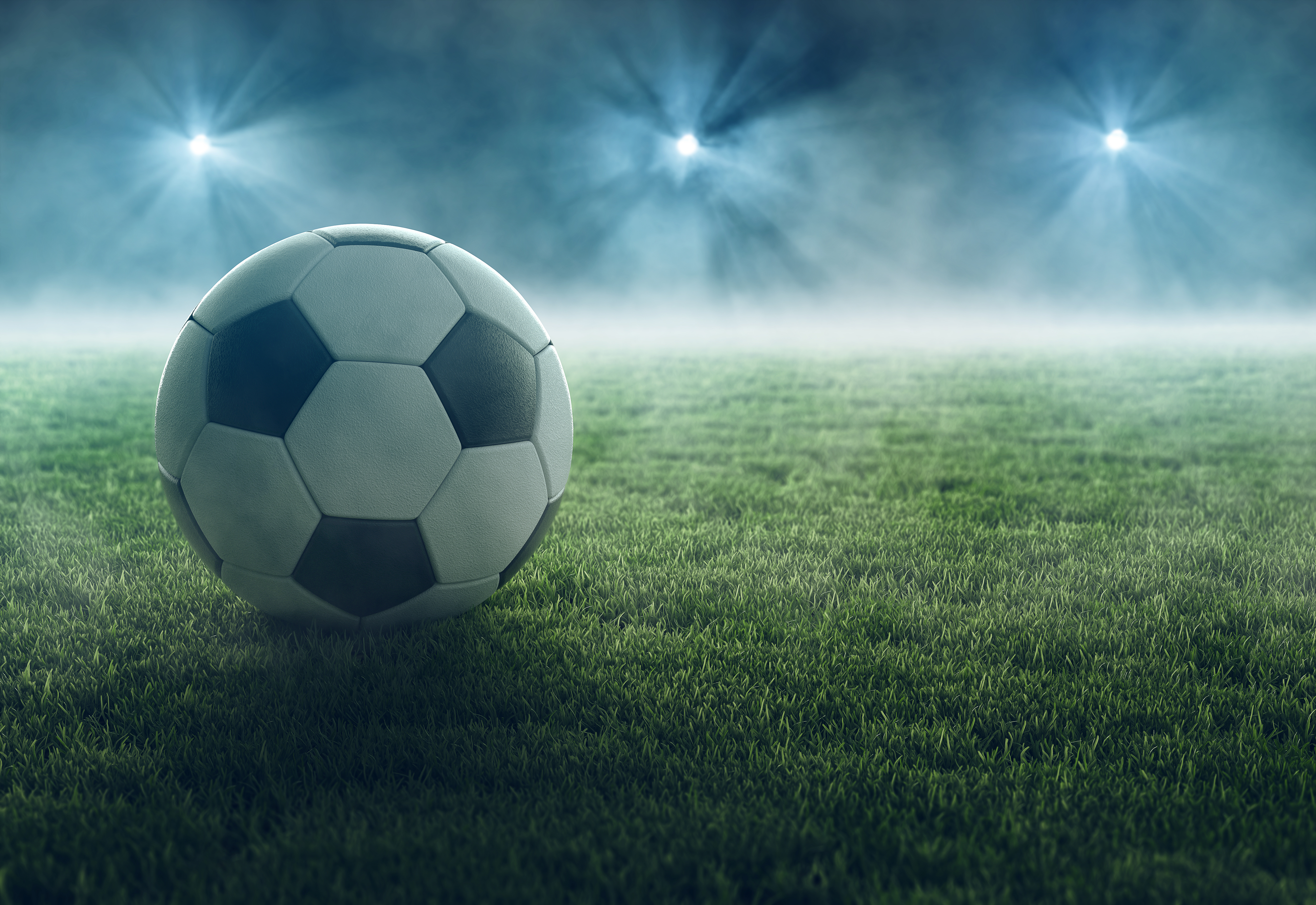 A black and white soccer ball rests on a lush green field. In the background, there is a soft mist, and bright lights illuminate the scene, creating an atmospheric effect. The setting suggests a nighttime sporting event.