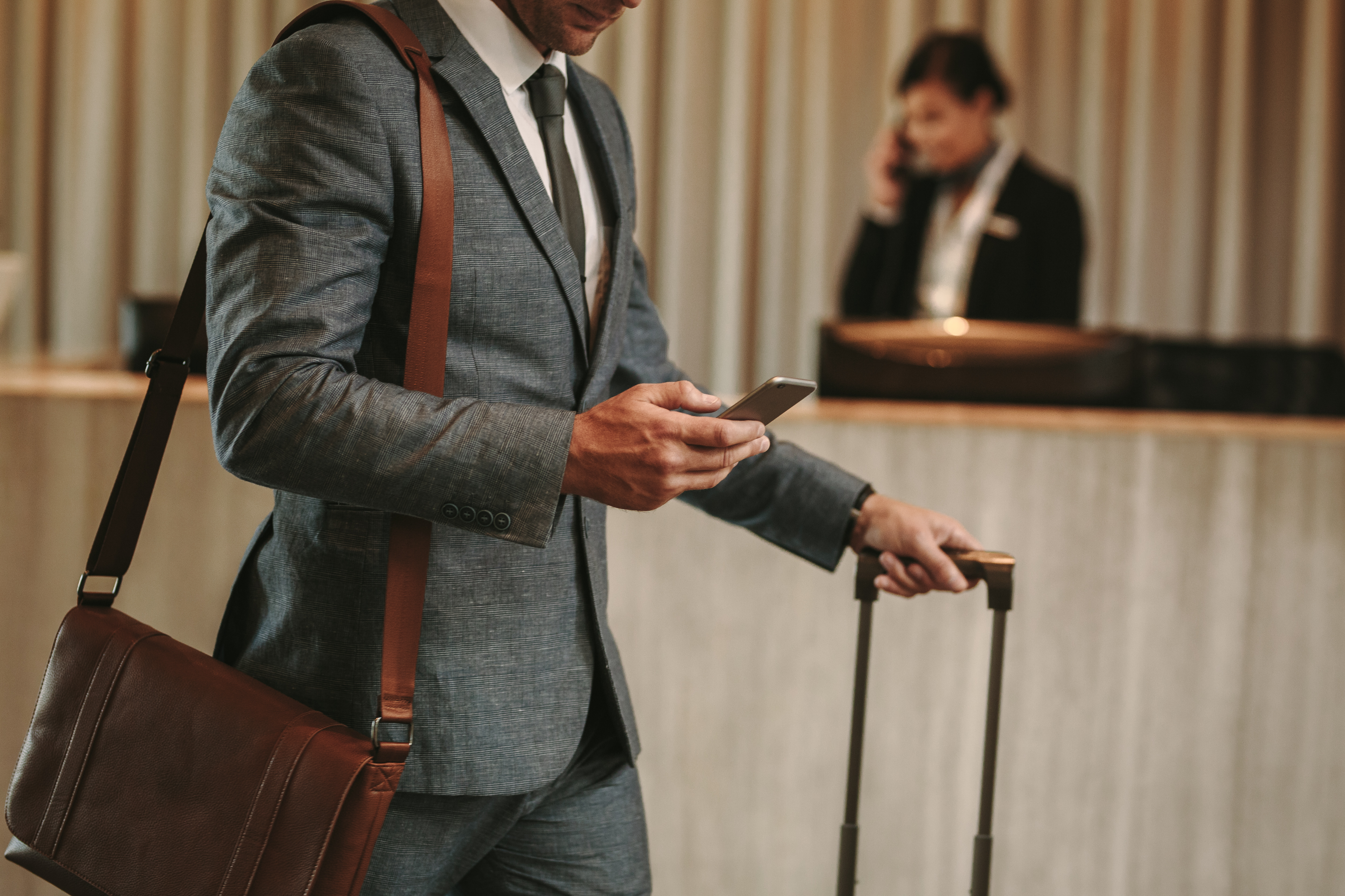 Ein Mann in einem grauen Anzug steht an einer Hotelrezeption. Er hält ein Smartphone in der Hand und hat eine Tasche über der Schulter. Neben ihm steht ein Koffer mit Teleskopgriff. Im Hintergrund ist eine Hotelmitarbeiterin am Telefon zu sehen.