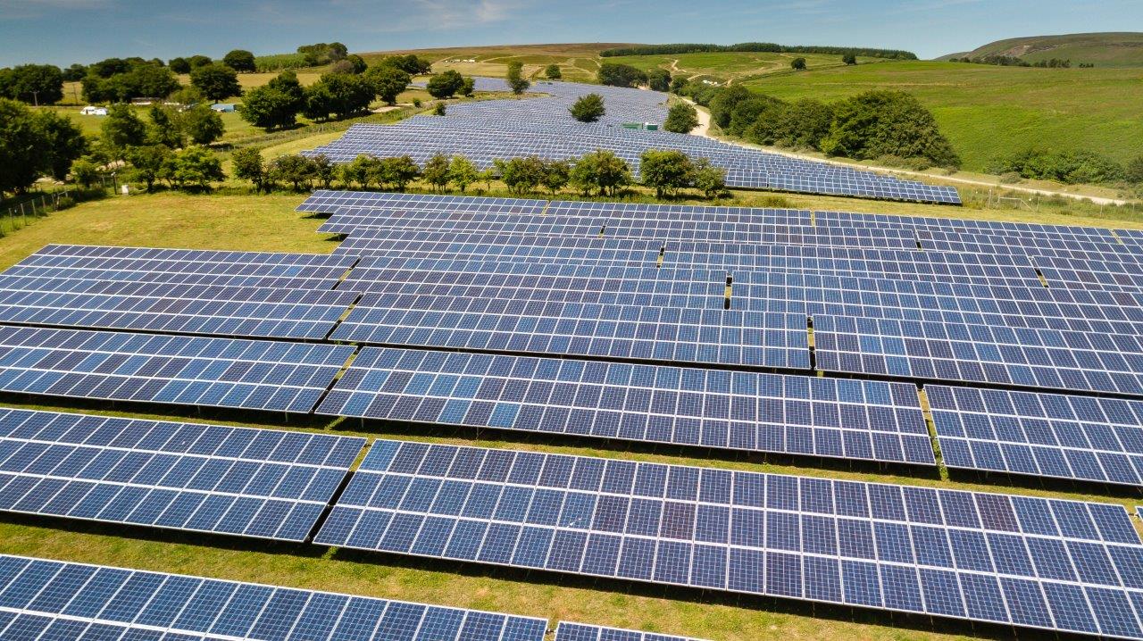 Aerial view of a large solar farm, featuring numerous rows of solar panels arranged neatly across a green field. The panels reflect sunlight, indicating energy production, while trees line the perimeter, with hills visible in the background.