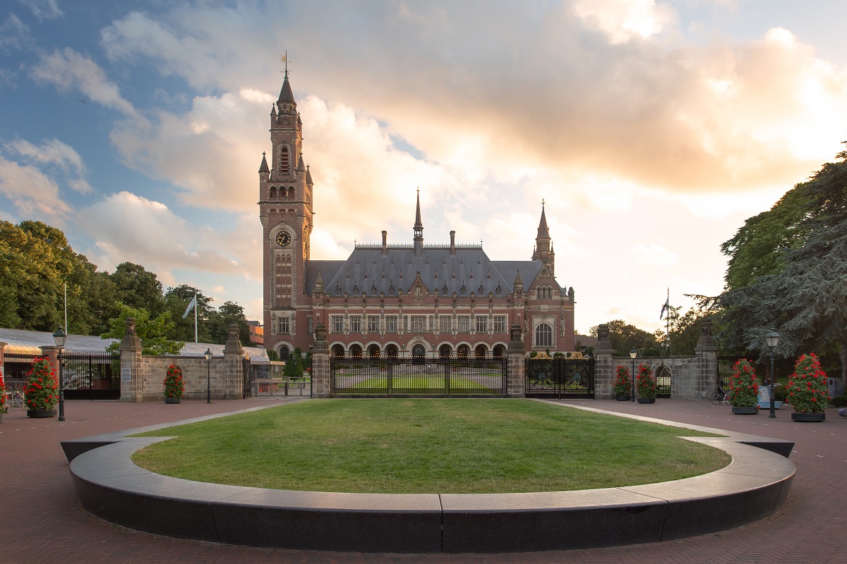 Ein historisches Gebäude mit einem markanten Uhrturm, umgeben von einem geschmackvollen Garten. Die Fassade ist reich verziert, und im Vordergrund liegt eine grüne Rasenfläche. Die Szene wird von einem bewölkten Himmel mit sanften Farben beleuchtet.