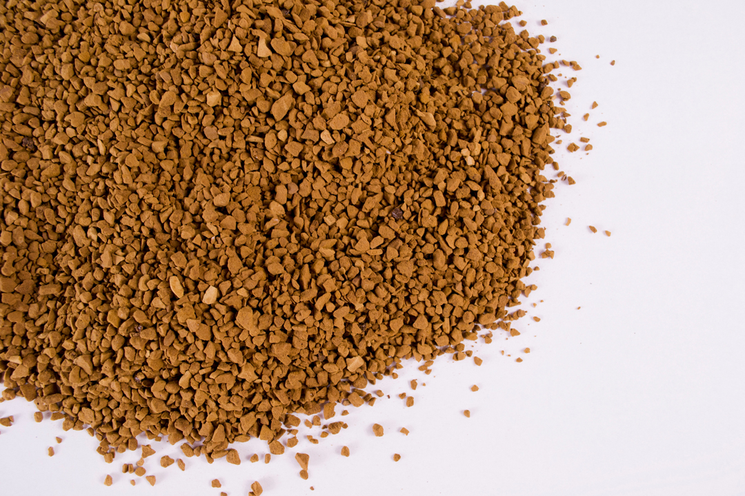 A mound of coarse coffee granules in shades of brown is scattered across a light surface. The texture appears rough and uneven, suggesting the granules are intended for brewing coffee.