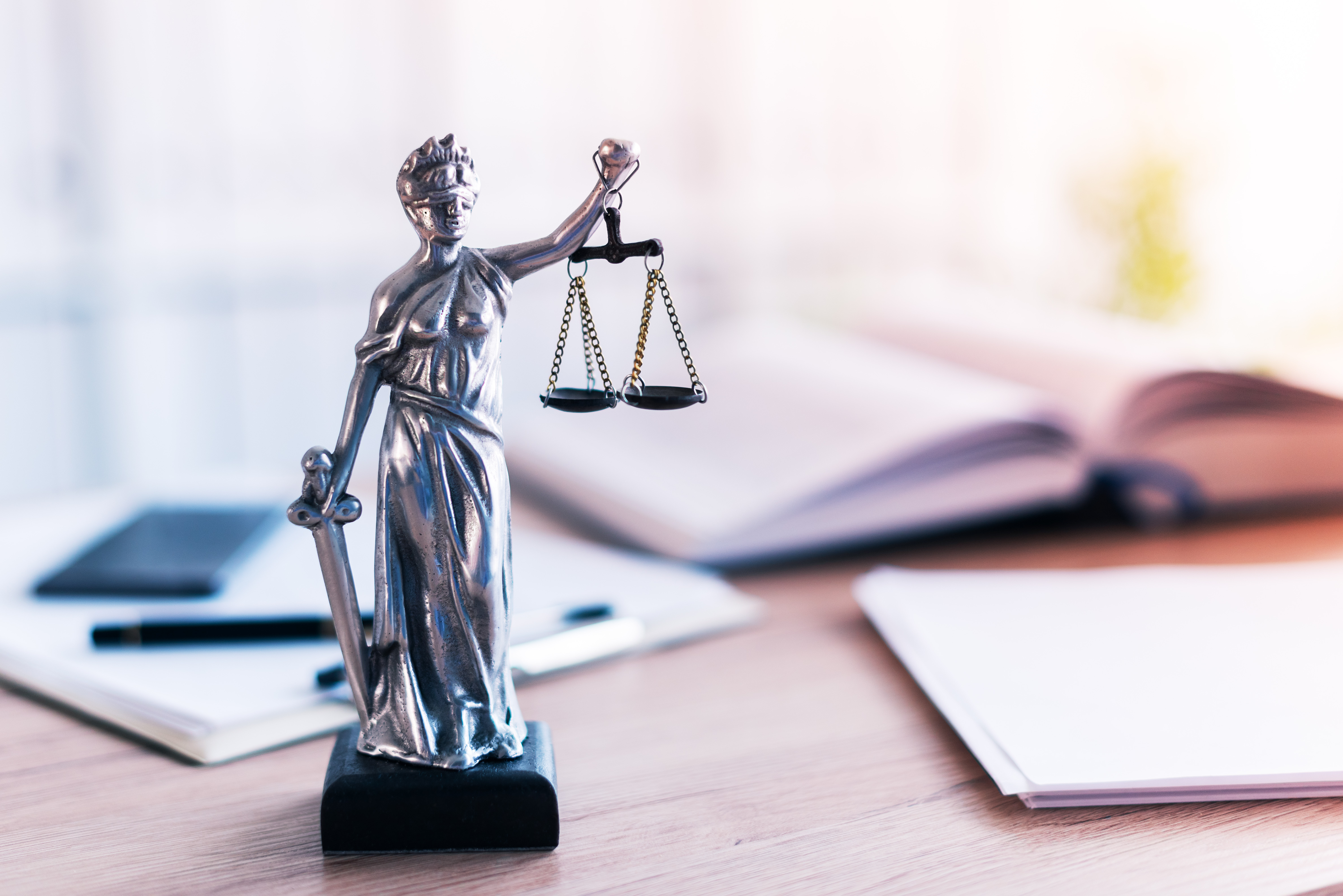 Eine kleine Statue der Justitia hält in einer Hand eine Waage und in der anderen ein Schwert. Im Hintergrund sind ein aufgeschlagenes Buch und Schreibutensilien zu sehen. Die Szene vermittelt das Thema Recht und Gerechtigkeit.