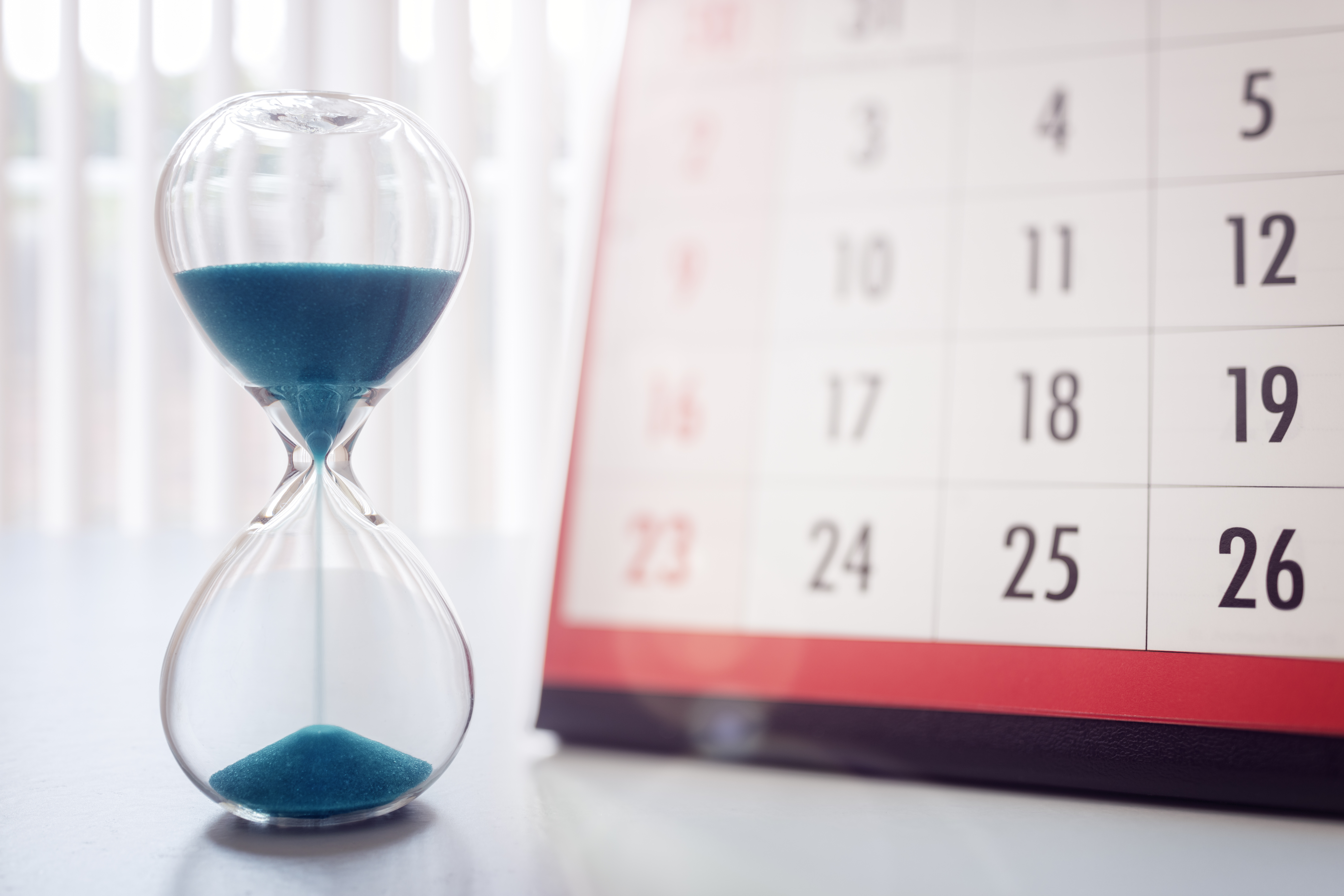 Eine Sanduhr mit blauen Sandkörnern steht auf einem Tisch, während im Hintergrund ein Kalender sichtbar ist. Die Uhr zeigt an, wie Zeit vergeht, und der Kalender zeigt verschiedene Tage des Monats an.